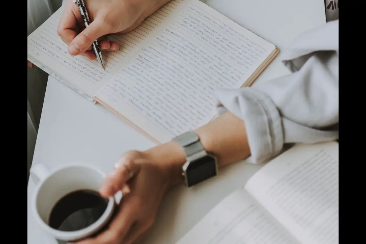 Hands writing in a notebook beside a cup of coffee and an open book